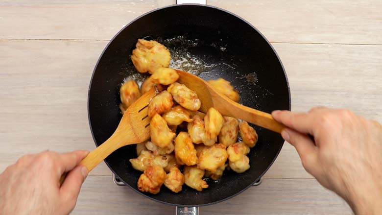 Hands holding two wooden spatula and stir frying batter fried chicken in honey sauce in a black wok – Honey Chicken Recipe