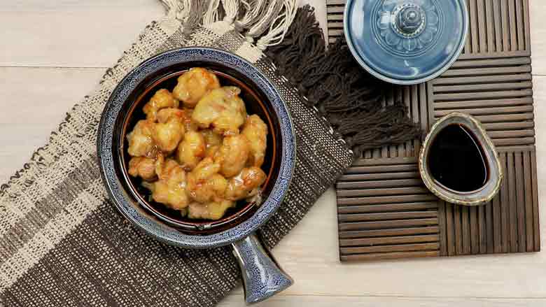 Honey chicken served in a single handled blue ceramic dish placed on a hand woven grey and white cotton mat with small bowls in the background – Honey Chicken