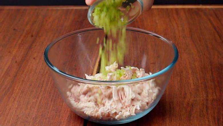 Diced celery being added to shredded chicken in a large glass bowl for chicken salad