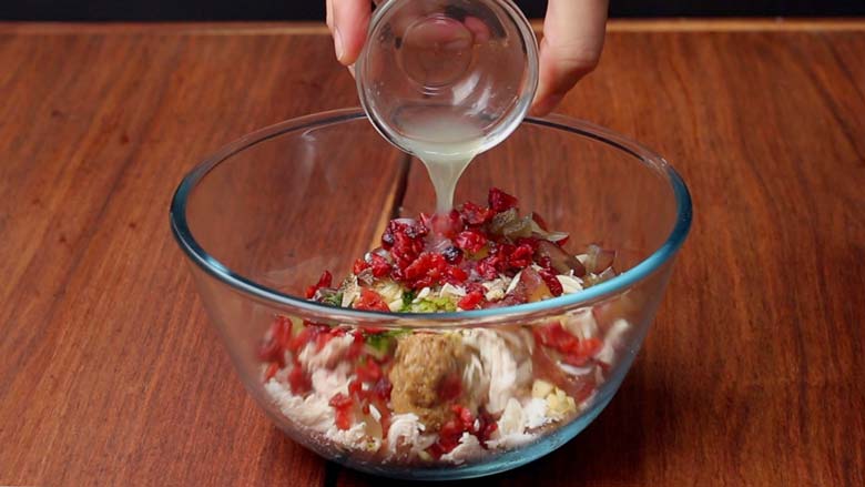 Lime juice being added to shredded chicken and other ingredients like cranberries, red grapes, mustard, pickled gherkins in a large glass bowl lying on a wooden table