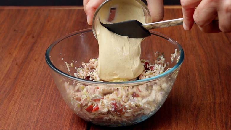 Using a rubber spatula to add homemade mayonnaise to all the other ingredients of the chicken salad in a glass bowl on a wooden table