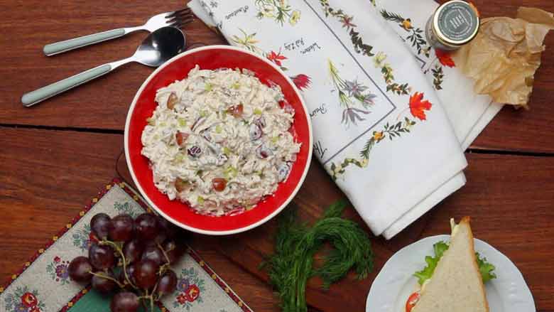 Chicken Salad plated in a red bowl with a white rim placed on a wooden table alongside red grapes, a cheese lettuce tomato sandwich, a printed cloth, a printed place mat, and a spoon and fork