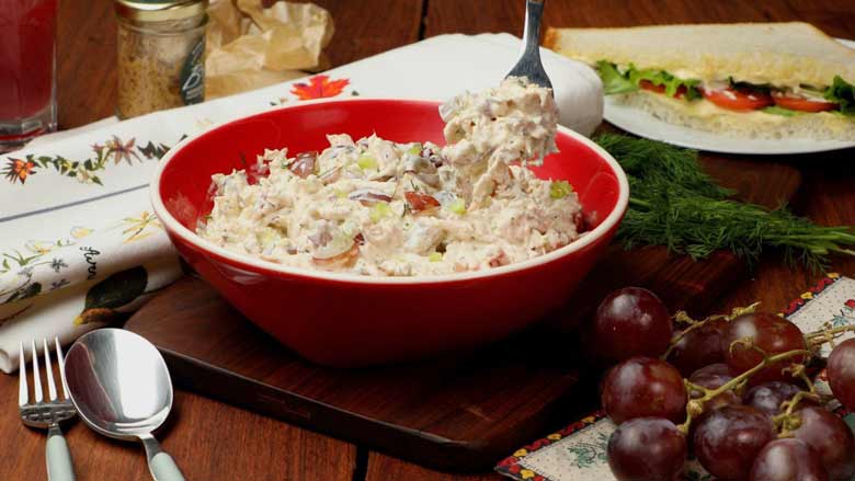 Serving chicken salad with a spoon from a red ceramic bowl with white rim with red grapes in the foreground and white printed cloth and sandwich in the background