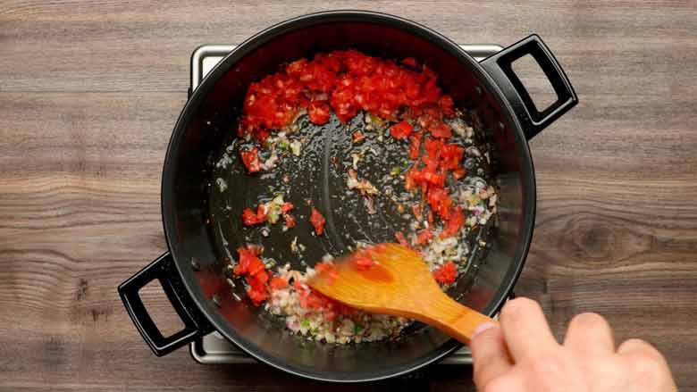 Stirring tomatoes with a wooden spatula along with fried onions in a black pan for the tadka