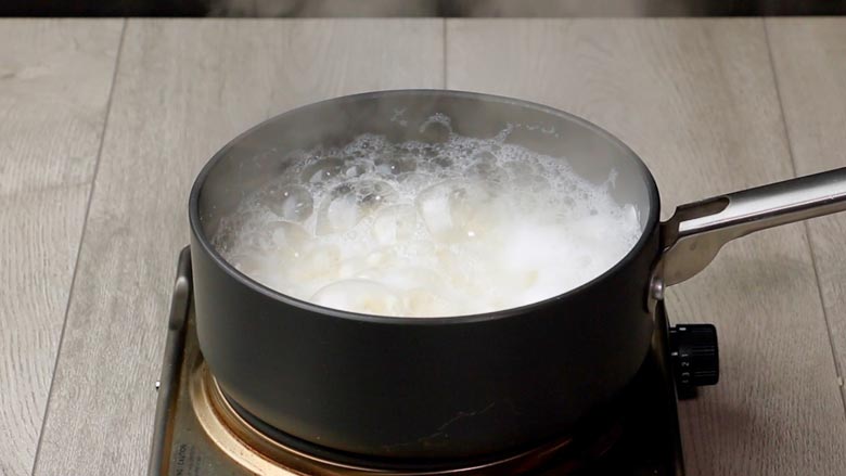 Raw rice being boiled in a black saucepan on a textured white wooden table in preparation for Lemon Rice