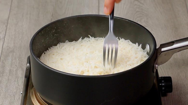 Using a fork to fluff cooked rice in a black pan on a white textured wooden table for the Lemon Rice recipe