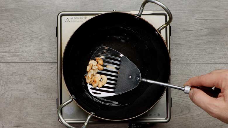 Golden brown cashew nuts being removed from a small black kadhai with a slotted steel spatula after being fried in oil for lemon Rice