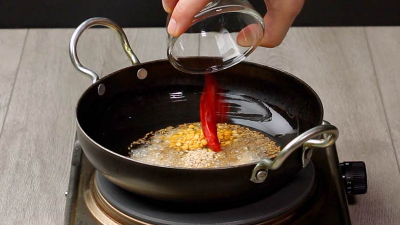 Dried red chillies being added from a small glass bowl to the dals and spices being fried in oil in a black kadhai as the tempering for Lemon Rice
