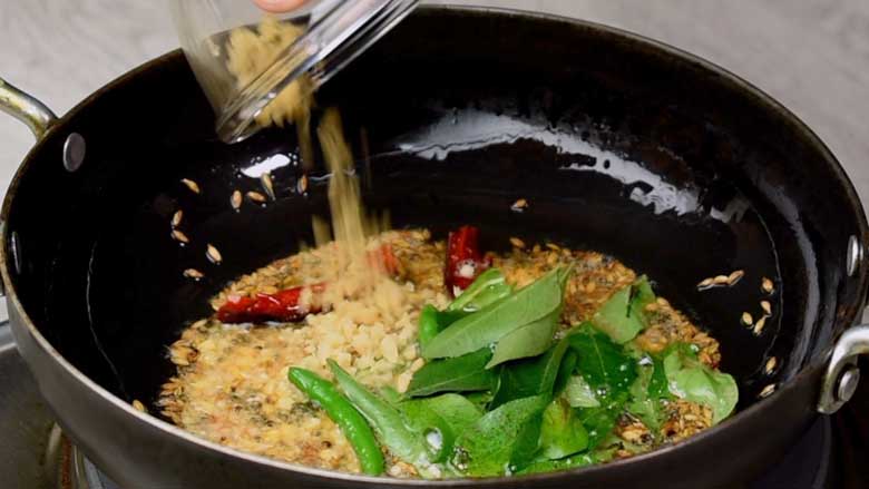 Chopped ginger being added to the tempering spices, dals, and curry leaves frying in oil in a black kadhai for lemon Rice