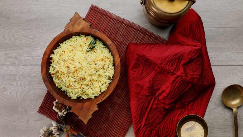 A top view of lemon Rice in an antique wooden bowl lying on a brick-coloured cane mat next to a red quilted silk cloth with a brass cup and large brass spoon.