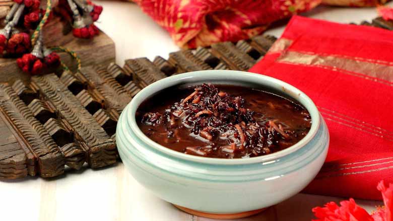 Kavanarisi - South India black rice breakfast dish in an aqua coloured ceramic bowl lying on a textured white table next to a carved wooden board and red and gold textiles, flowers, and tassles