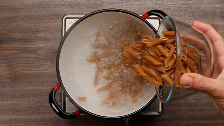 Whole wheat pasta - penne - being tipped from a glass bowl into salted boiling water in a black rimmed white enamel pot on an electric stove