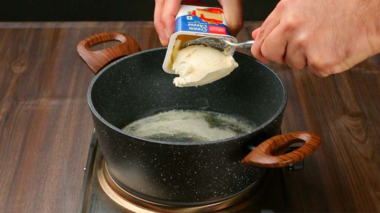 Spooning cream cheese from a plastic container into melted butter in a black and white speckled pan with two wooden handles