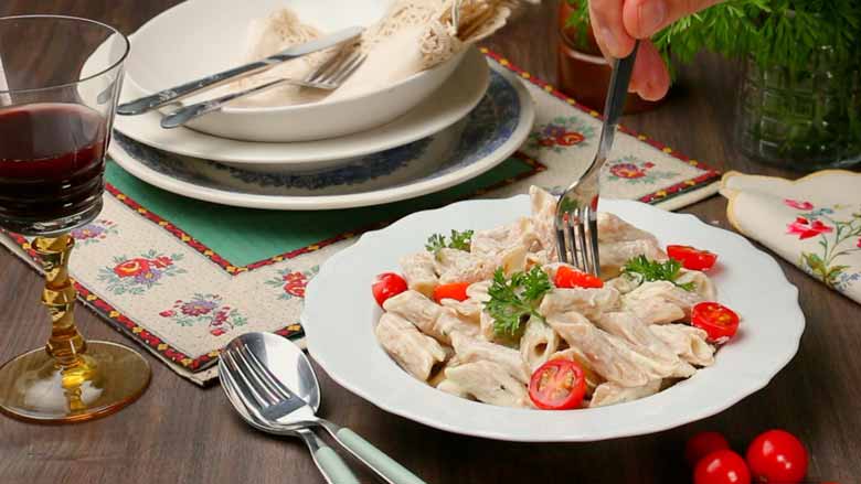 Using a fork to pick up white sauce pasta with cheese with cherry tomatoes and parsley from a shallow white pasta bowl lying on a textured wooden table with a glass of wine lying next to it