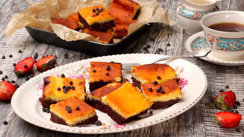 Cheesecake Brownies squares served on a floral bone china platter on a textured black and white wooden table. Placed next to a cup of tea, strawberries, and scattered chocolate chips.