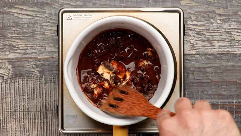 Butter and chocolate in a white rimmed pan with a yellow handle being stirred by a wooden spatula till melted. On a textured grey and white wooden table.