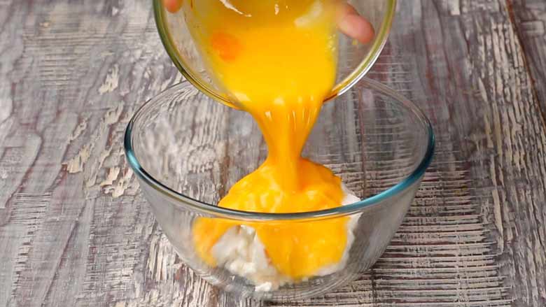 Lightly beaten eggs being poured over cream cheese in a glass bowl on a white and grey textured table to make the cheesecake mixture for cheesecake brownies