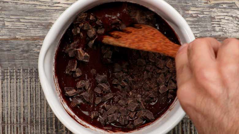 Chocolate Chips being folded into the chocolate brownie batter in a white rimmed saucepan on a wooden grey and white textured table with a wooden spatula