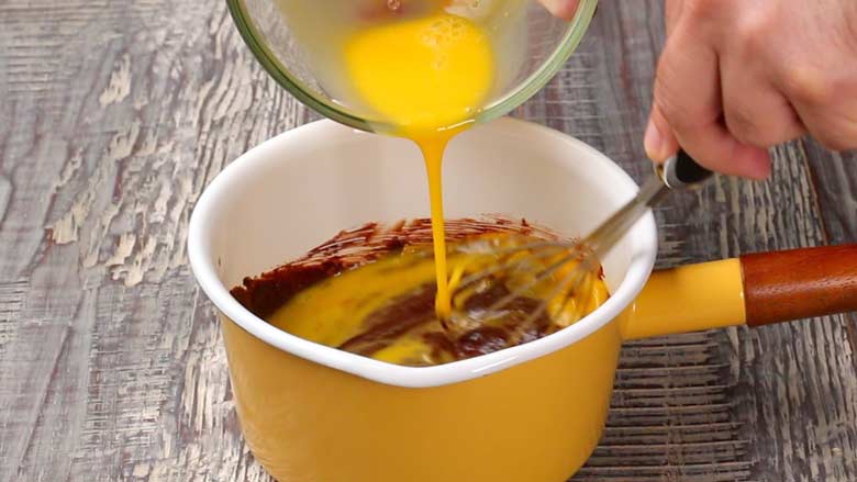 Lightly beaten eggs being whisked into a slightly cooled chocolate mixture in a yellow saucepan with a wooden handle off the heat on a textured wooden table.