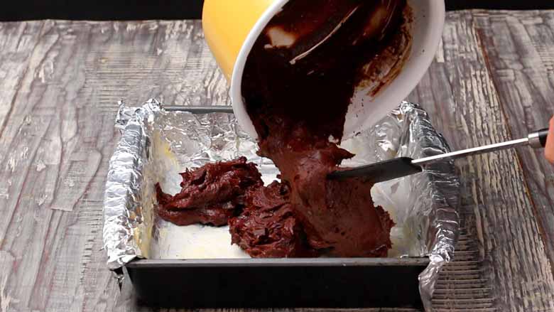 Brownie batter being tipped into a baking tin lined with foil from a yellow saucepan to make the bottom layer of Cheesecake Brownies
