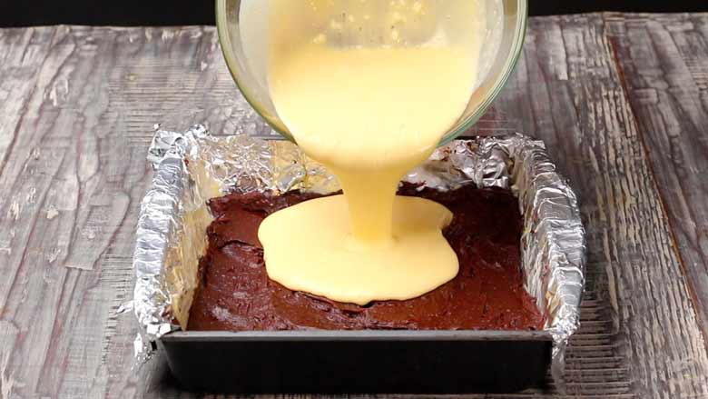 Pouring cheesecake batter from a glass bowl over the brownie batter in a baking tin prepared and lined with foil for Cheesecake Brownies