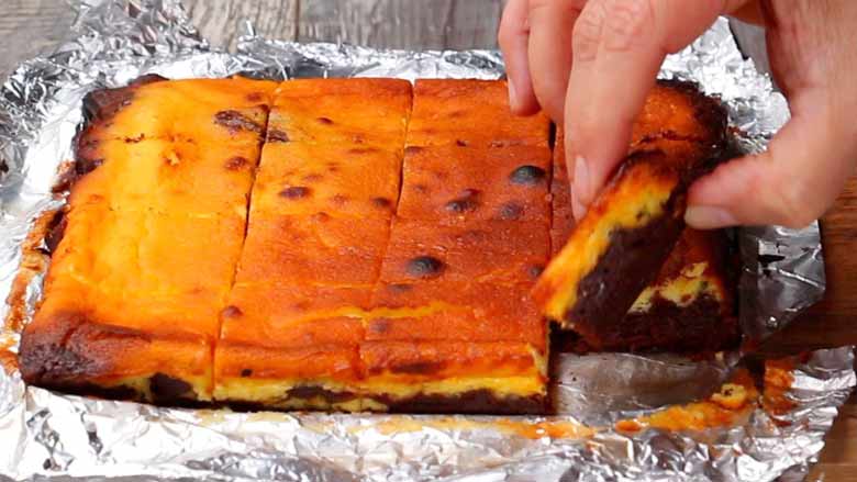 A hand picking up one freshly baked Cheesecake Brownie square from among the 16 squares on a sheet of foil