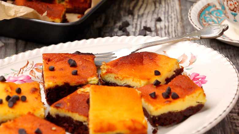 A closeup shot of Cheesecake Brownies garnished with chocolate chips served on a floral bone china plate with a fork lying on it and a baking tin with brownies and a cup and saucer in the background