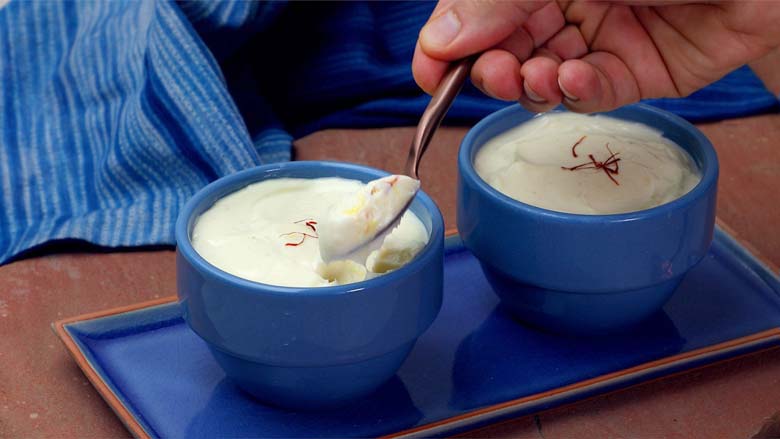 A hand using a spoon to pick up Saffron Shrikhand garnished with saffron strands in two blue ceramic bowls on a blue ceramic platter plate on a pink Jaipur stone with blue fabric in the background