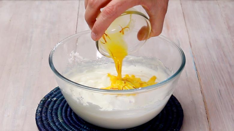 Saffron soaked in warm milk being poured into the whipped hung curd in a glass bowl on a blue coiled round mat for shrikhand