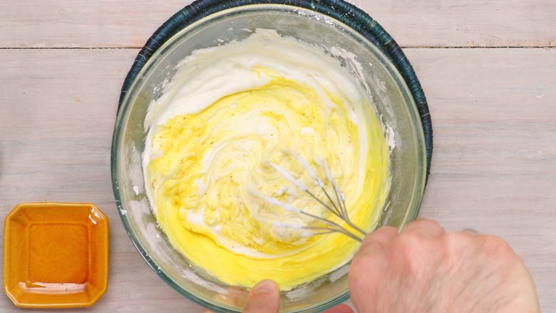 Saffron milk being whisked into the sweetened chakka or hung curd in a large glass bowl on a white textured wooden table for saffron shrikhand