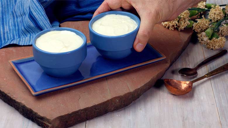 A hand placing two blue ceramic bowls filled with saffron shrikhand on a blue ceramic platter lying on a pink Jaipur stone with a blue fabric in the background
