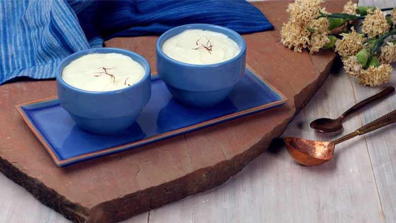 Saffron Shrikhand garnished with saffron strands in two blue ceramic bowls on a blue ceramic platter plate on a pink Jaipur stone with white flowers in the background