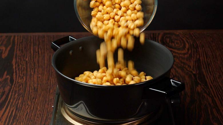 Soaked Chickpeas being tipped into a black pan on an electric stove lying on a textured wooden table