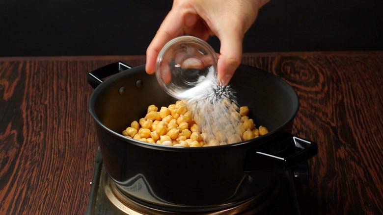 Baking soda being added from a small glass bowl to chickpeas cooking in a black pan for hummus