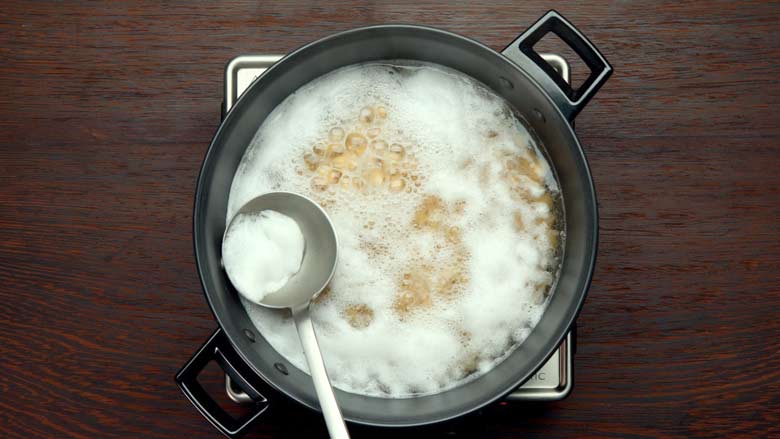 A metal ladle being used to skim foam and scum from boiling chickpeas in a black pan on an electric steel stove on a wooden table while making hummus