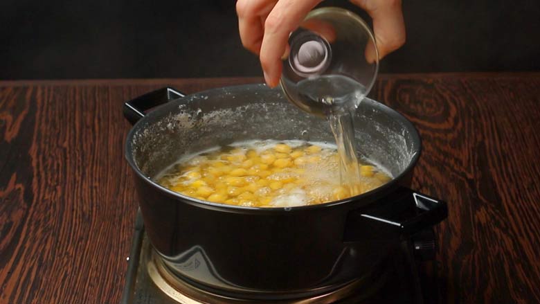 Cold water being poured from a glass bowl into the boiling chickpeas in a black pan while making homemade hummus