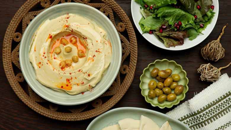  Top view of hummus garnished with chickpeas, olive oil, and red pepper powder in a green bowl on a cane bowl lying next to marinated olives, khobz bread, and a fresh green salad
