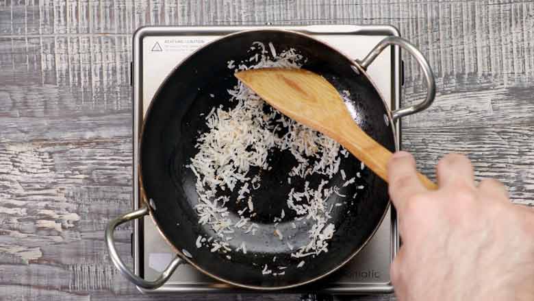 Top view of shredded dried coconut being dry roasted till golden in a black kadhai pan with steel handles using a wooden spatula on a steel electric stove to make Andhra Gunpowder 
