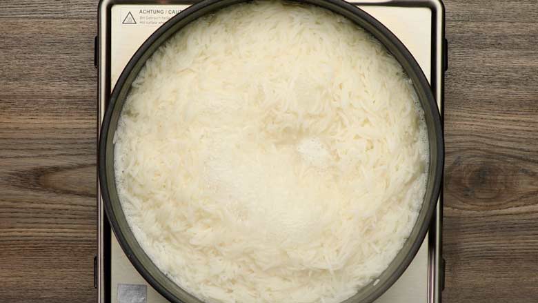 Rice boiling in salted water in a black saucepan on a steel electric stove lying on a wooden table in preparation for coconut rice