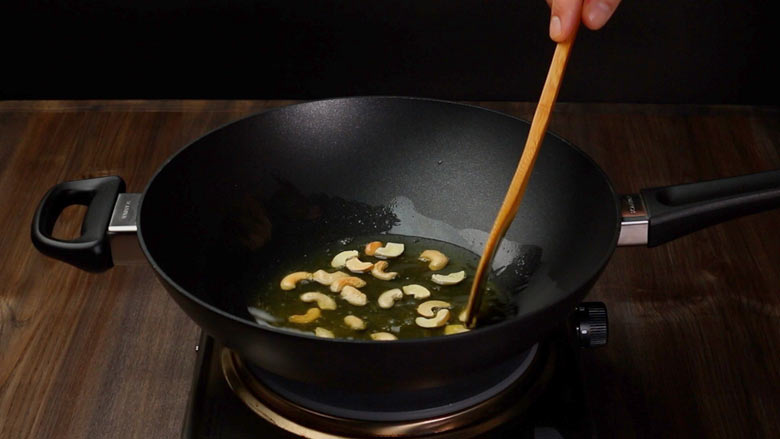 Cashew nuts being fried till golden in a black non stick wok stirred by a wooden spatula in preparation to be added to coconut rice