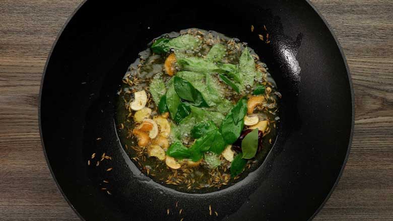Cashew nuts, mustard seeds, cumin seeds, dried red chilli, asafoetida (hing), curry leaves, and green chillies being fried in ghee in a black kadhai or wok on a wooden table for coconut rice