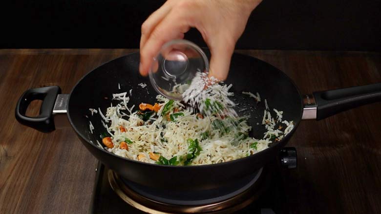 Salt being added from a small glass bowl to spices and coconut being sautéed in a black kadhai with a black handle for coconut rice