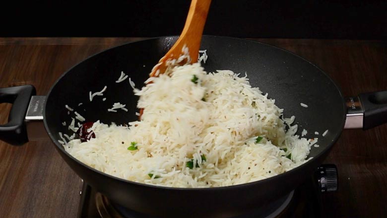 Cooked rice being gently mixed into sautéed spices and coconut in a black kadhai with a black handle on a wooden table for South Indian Coconut Rice