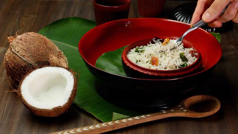 A hand holding a spoon serving South Indian Coconut Rice from a red and black Burmese lacquer bowl on a banana leaf with a cut coconut and a wooden inlaid spoon in the foreground and another black Burmese lacquer container in the background