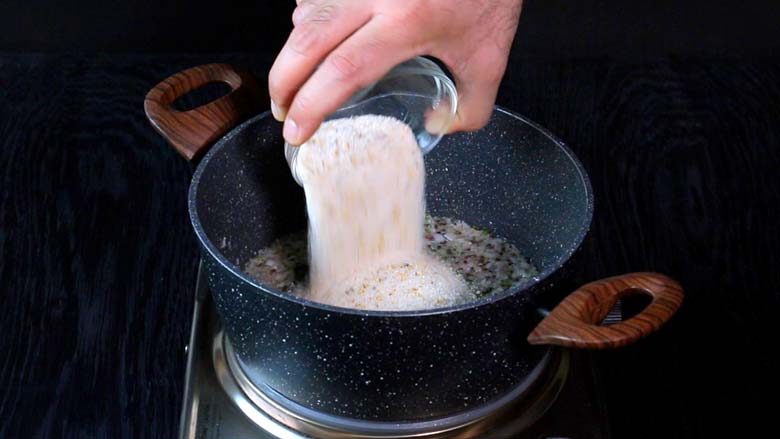 Coarsely ground rice and spices being added from a small glass bowl to the prepared tempering in a grey and white speckled pan with wooden handles for rice upma