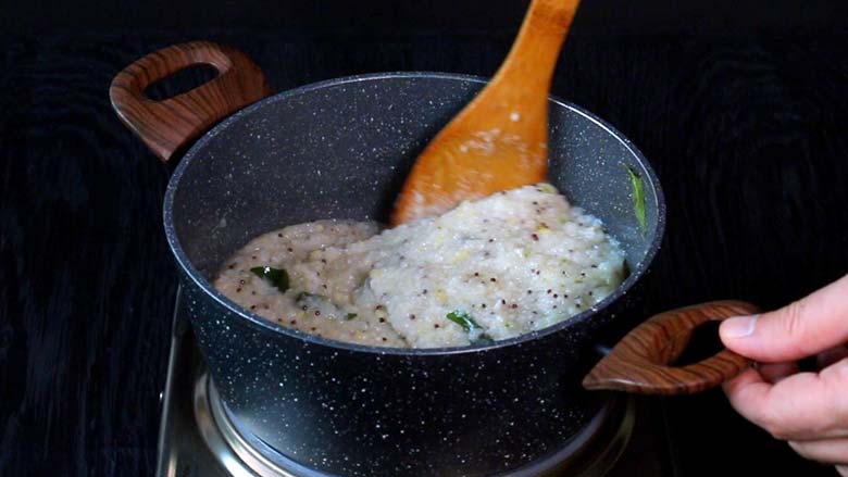 Cooking and stirring the rice upma with a wooden spatula till it thickens in a grey and white speckled pan with wooden handles on an electric stove - rice upma recipe