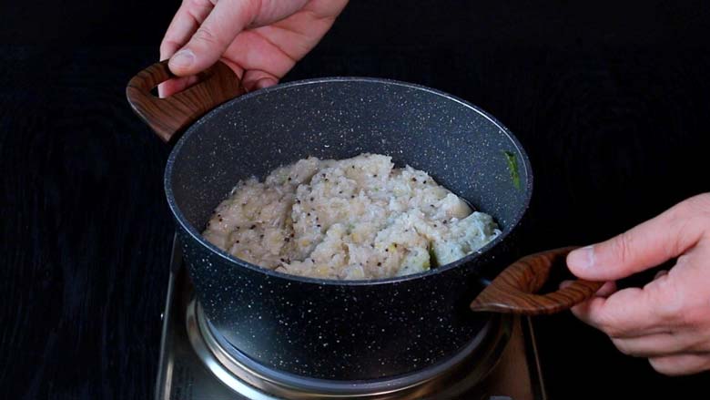 Two hands taking the cooked rice upma in grey and white speckled pan with wooden handles off the electric stove