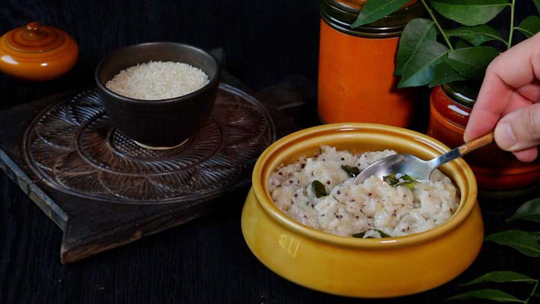 Rice Upma being served with a metal spoon from a ochre ceramic dish. A lacquered brick-coloured bowl and hand carved wooden platter with a small bowl with rice lies the background - Rice Upma Arisi Upma Recipe