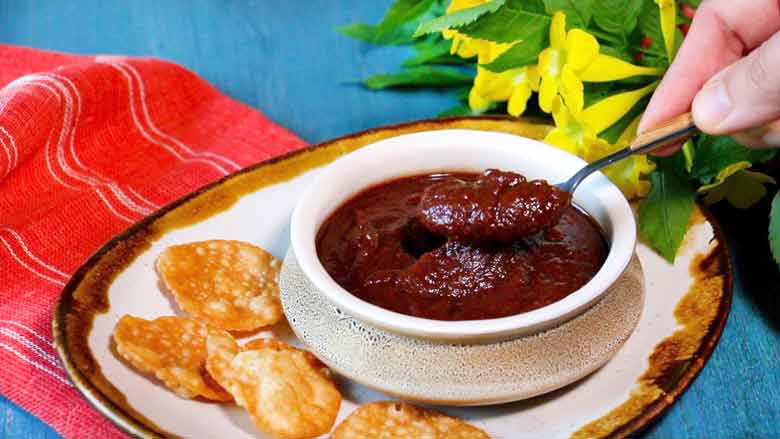 Date and Tamarind Chutney being served from a ceramic bowl lying on a ceramic platter on a blue table with a red cotton cloth and yellow flowers in the background - Sweet Chutney Recipe