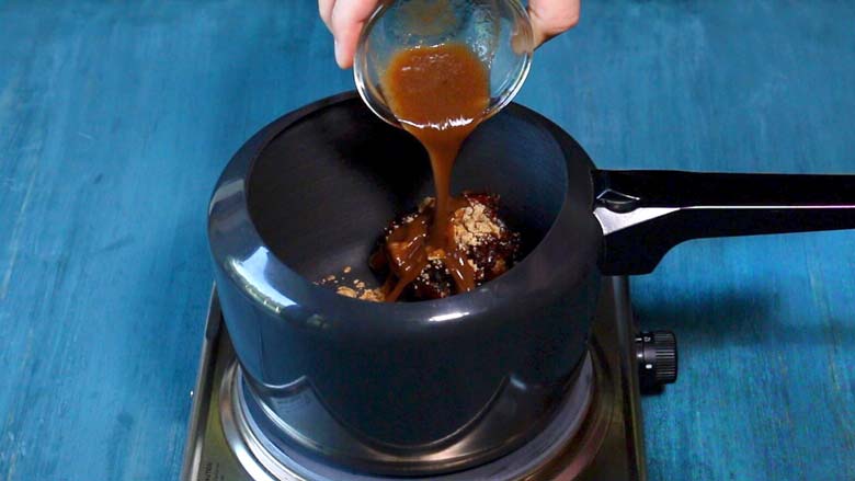 Tamarind paste being poured from a small glass bowl into a black pressure cooker with tamarind and jaggery in it.  This rests on a metallic electric stove on a blue table - Sweet Chutney Recipe